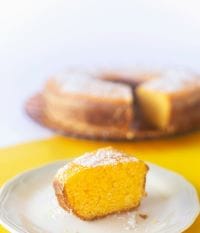 A a single slice of Gari cake dusted with powdered sugar on a white plate, with the rest of the ring-shaped cake blurred in the background against a yellow and white backdrop.