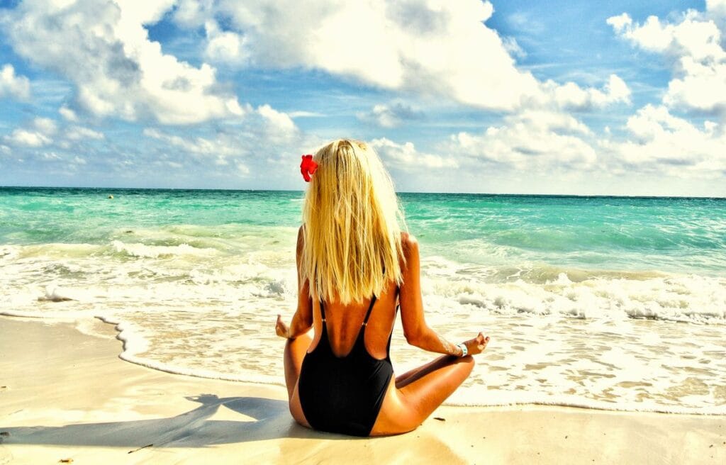 A blonde lady meditates on the white sand beach at The Ultimate Haven Hotel on the private THEBI Island in the Bahamas, overlooking turquoise Caribbean waters.